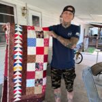 A man holding up a quilt on the porch.