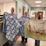 Three people holding colorful quilts indoors.
